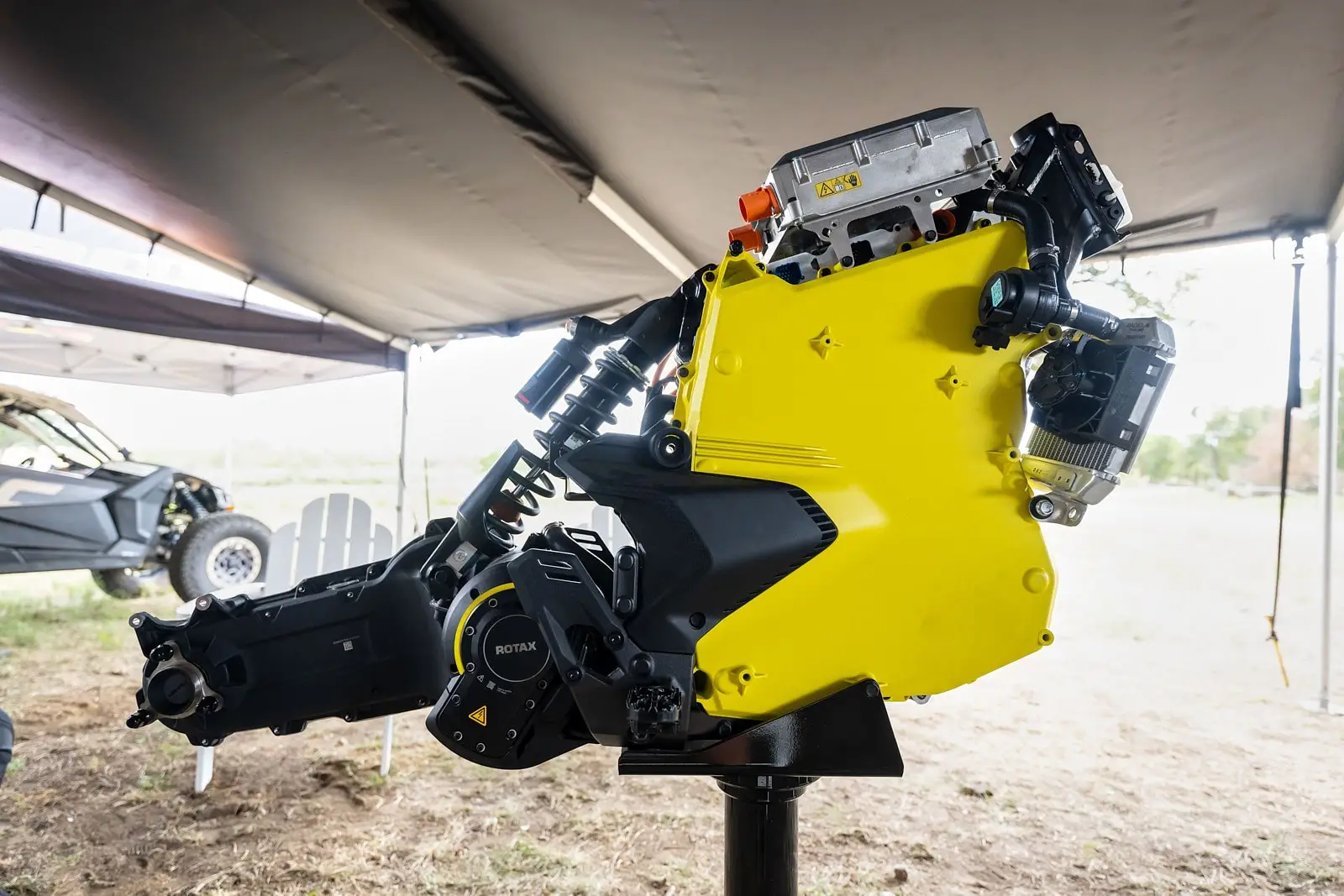 Exposed yellow electric motor block mounted on a stand, showing visible suspension and components, partially surrounded by an outdoor tent.