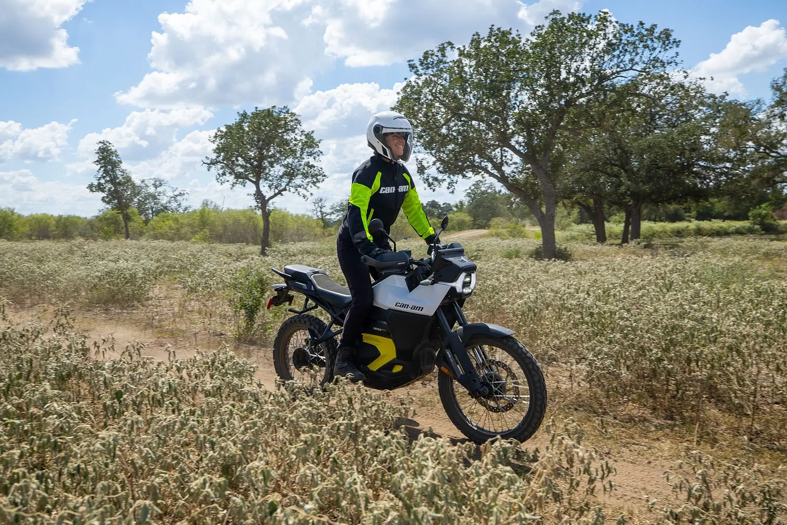 Person wearing a helmet rides on a Can-Am Origin electric motorcycle pulse on dirt road surrounded by sparse vegetation and trees.