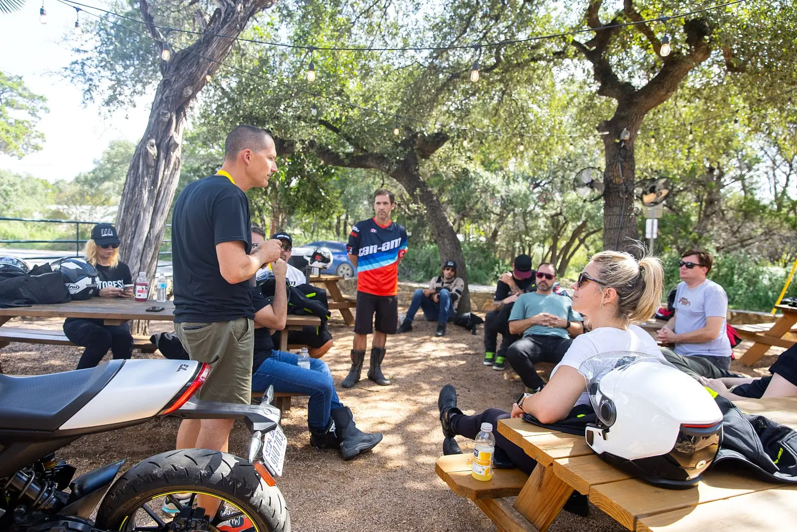 A group of people seated outdoors, listening to a speaker. Many are wearing motorcycle gear, and helmets are placed on surrounding picnic tables.