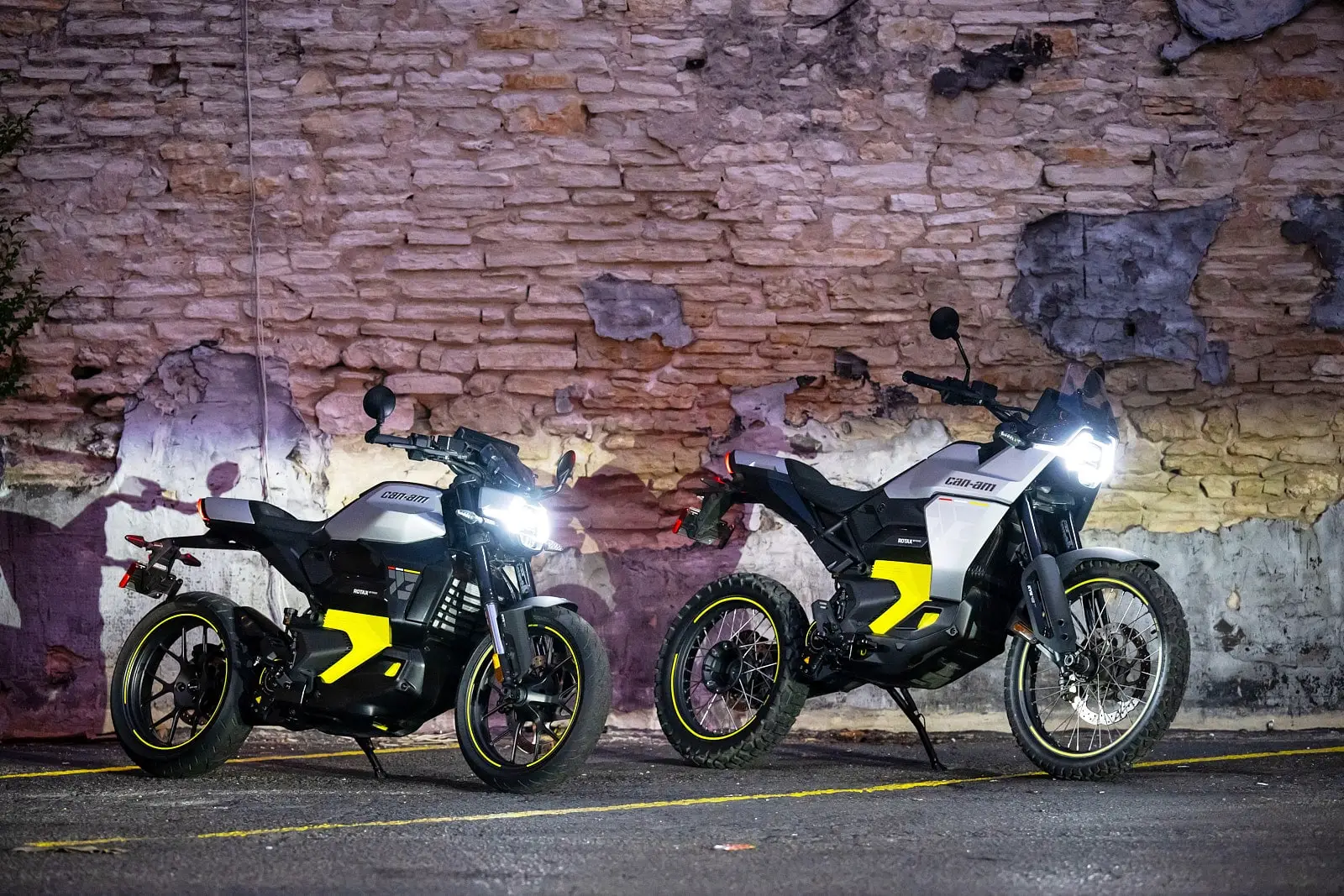 Two electric motorcycles from Can-Am, models Pulse and Origin, are parked side by side in front of a brick wall illuminated.