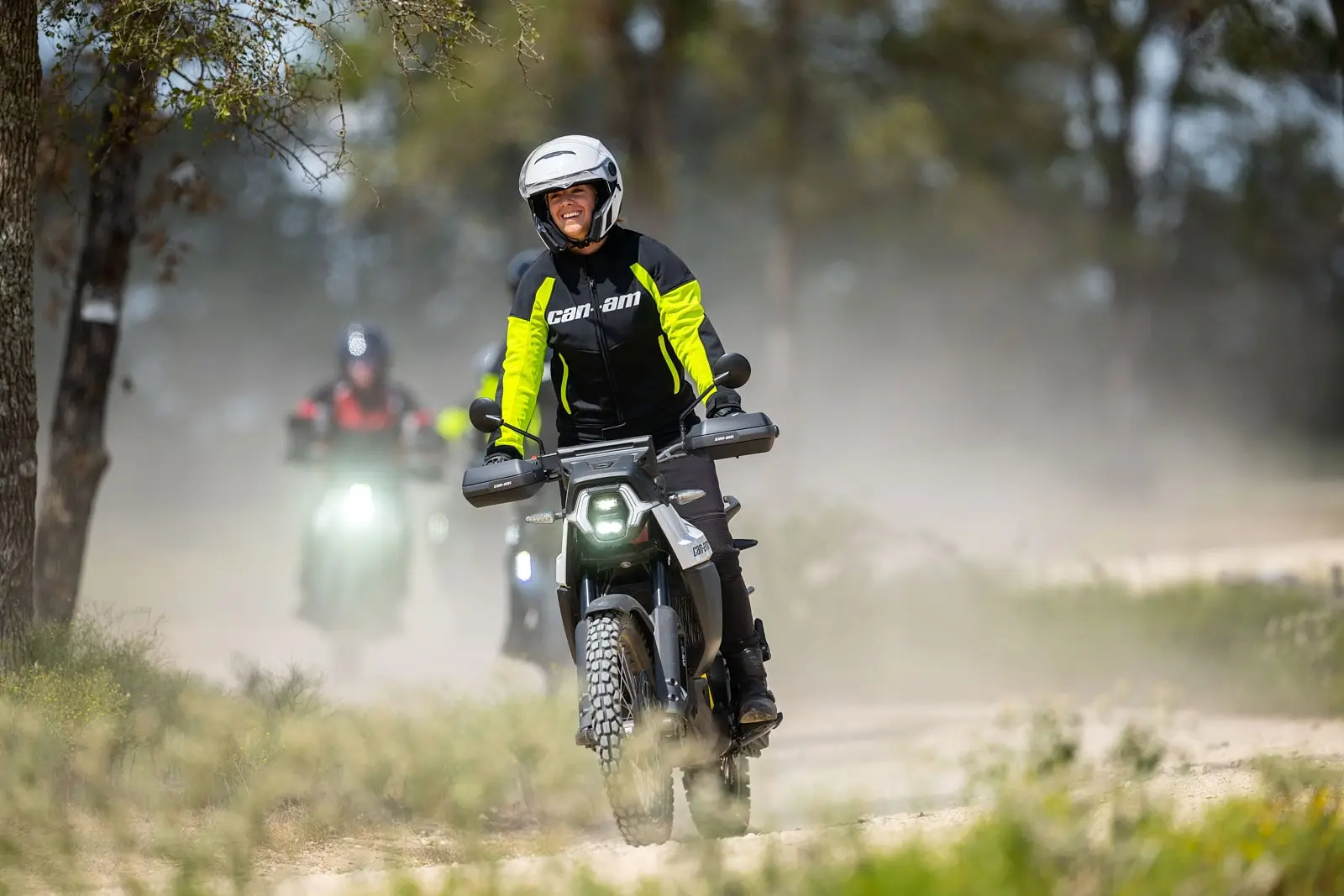 Un groupe de personnes roule sur des motos Can-Am Origin, soulevant de la poussière sur un chemin de terre entouré de verdure. La personne en tête affiche un grand sourire.