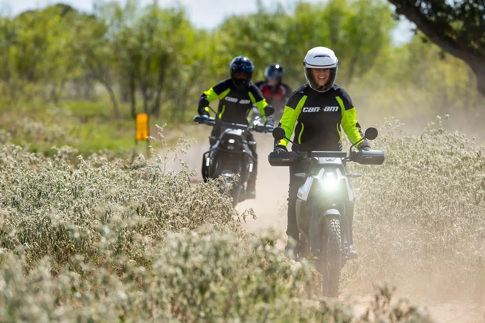 Groupe de motards en motos électriques Origin roulant sur un sentier poussiéreux entouré de végétation, avec le pilote de tête souriant.