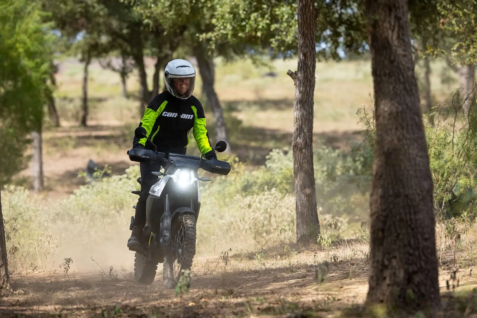 A smiling rider on a Can-Am Origin motorcycle navigating a forest trail surrounded by trees and vegetation.