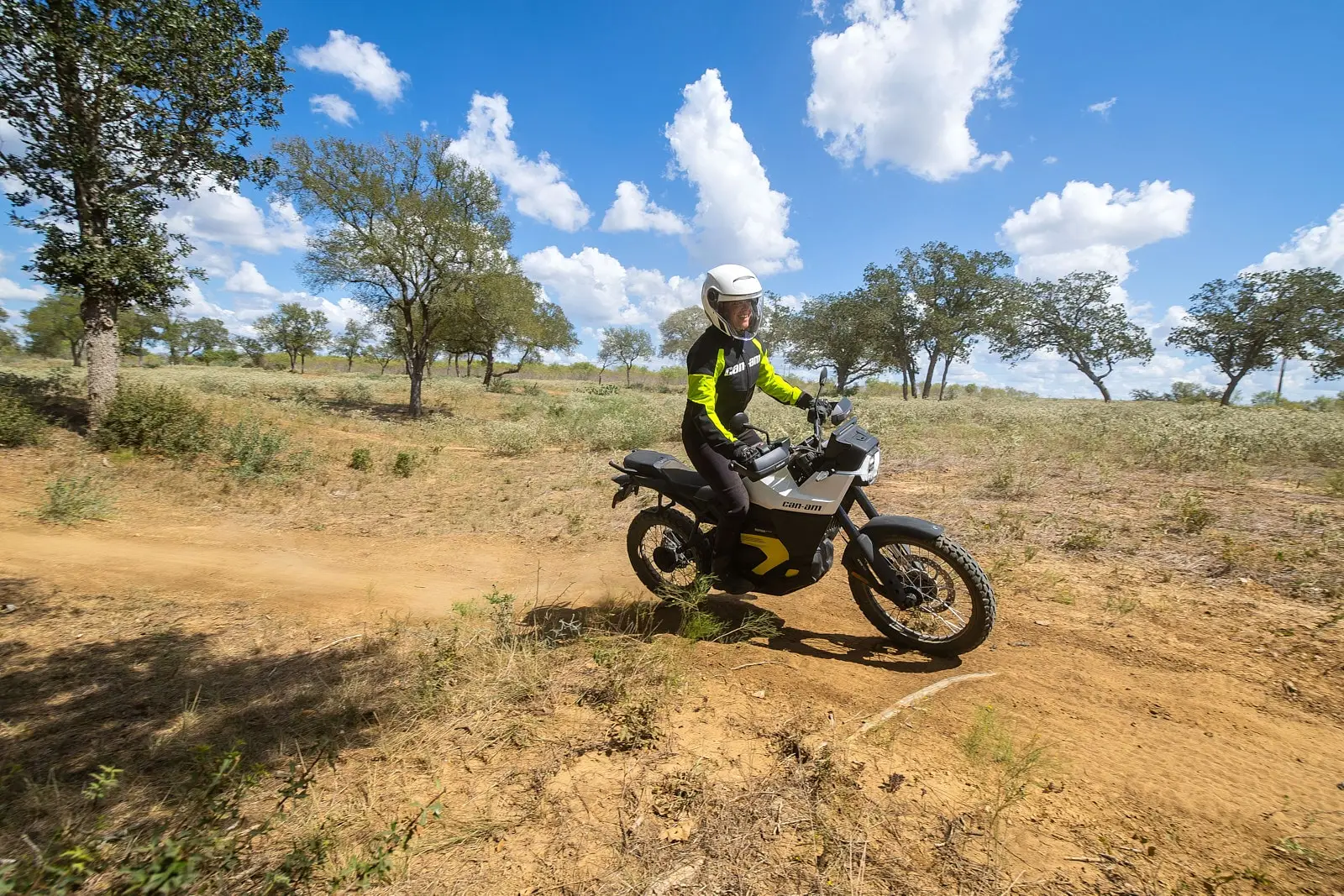 Motard en tenue Can-Am pilotant une moto électrique sur un sentier poussiéreux, sous un ciel bleu avec des nuages et entouré de végétation clairsemée.