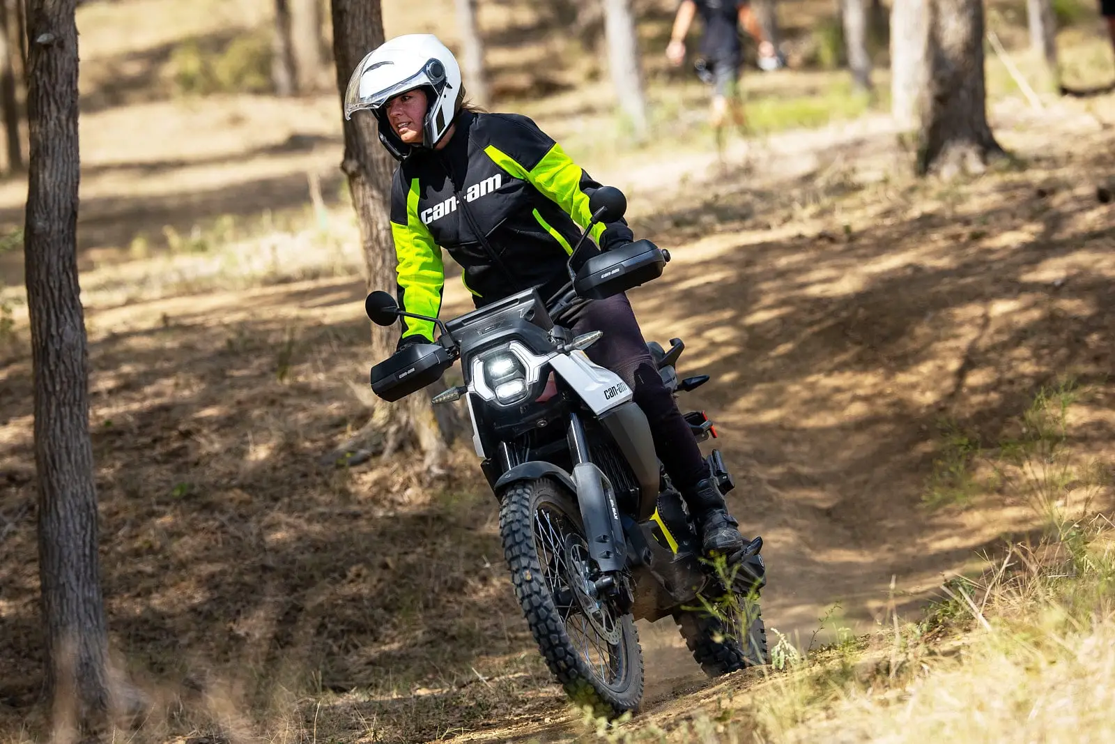 A rider on a Can-Am electric motorcycle on a forest trail, wearing Can-Am safety gear and a white helmet.