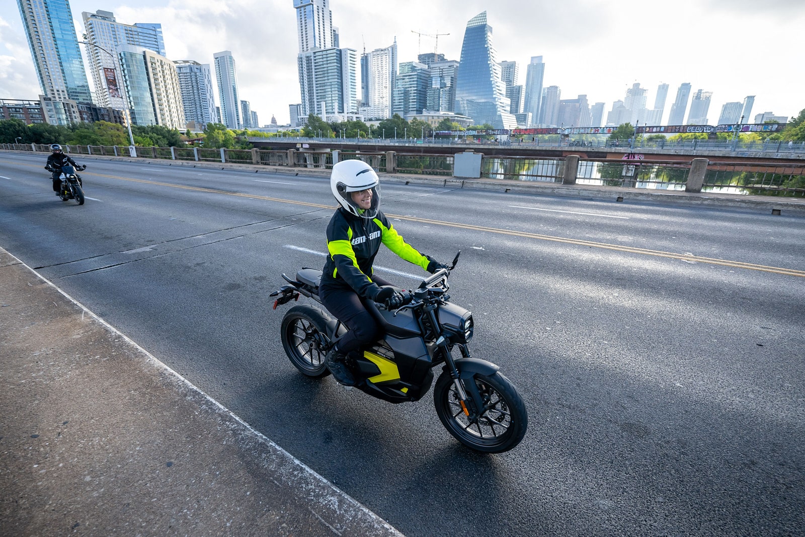 Une personne conduit une moto Can-Am électrique sur un pont, avec la silhouette de gratte-ciels en arrière-plan.