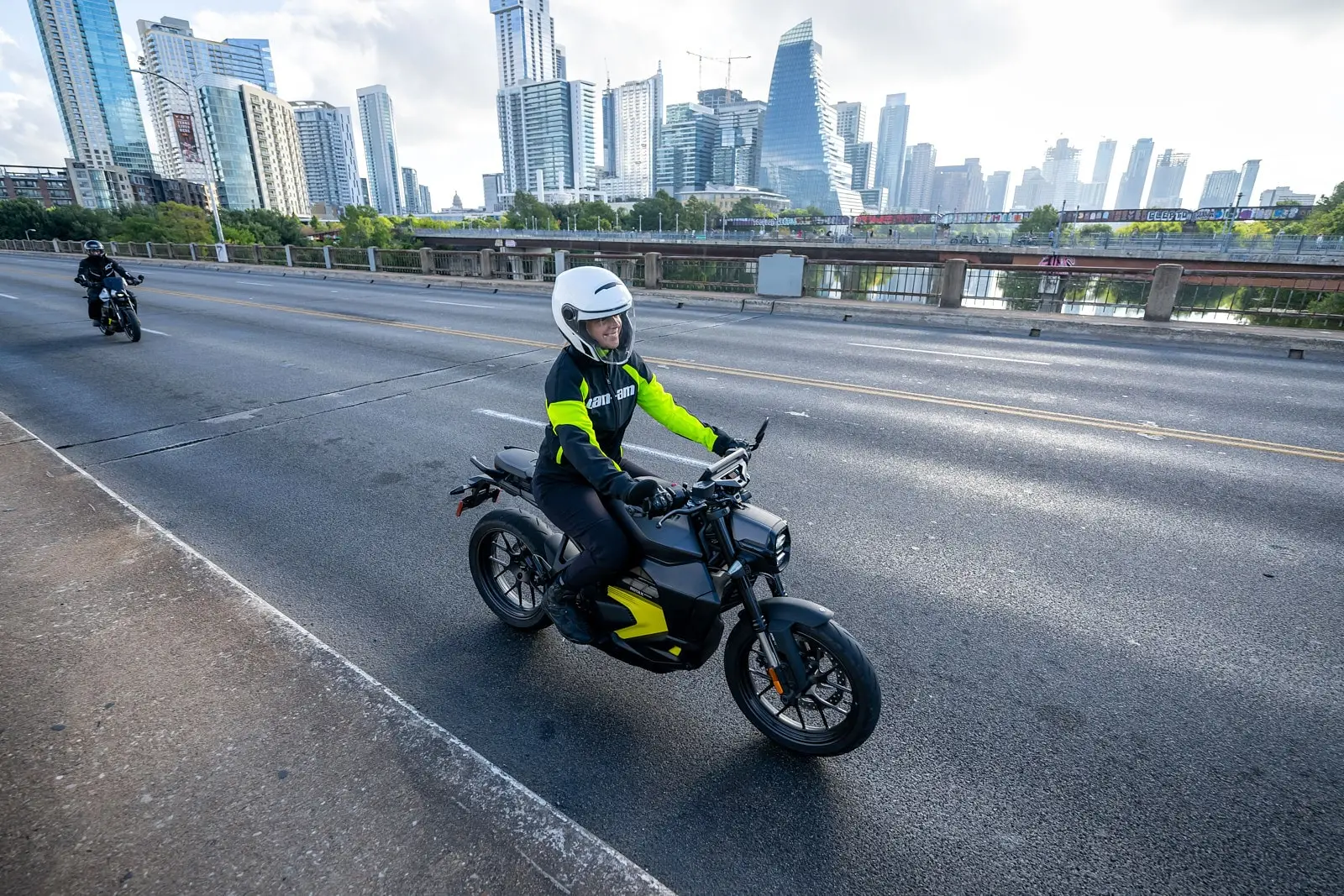 A rider on a Can-Am electric motorcycle crosses a bridge with a city skyline in the background.