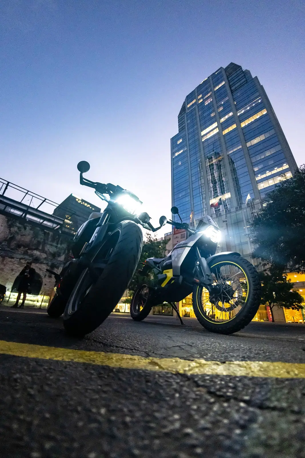 Two Can-Am electric motorcycles parked from a low-angle view at dusk, with a tall glass building in the background.