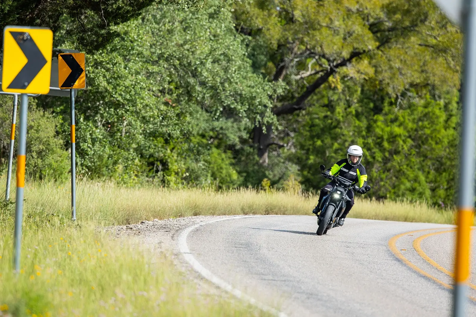 Un motard sur une moto Can-Am Pulse prend un virage sur une route bordée de panneaux de signalisation et d’arbres.