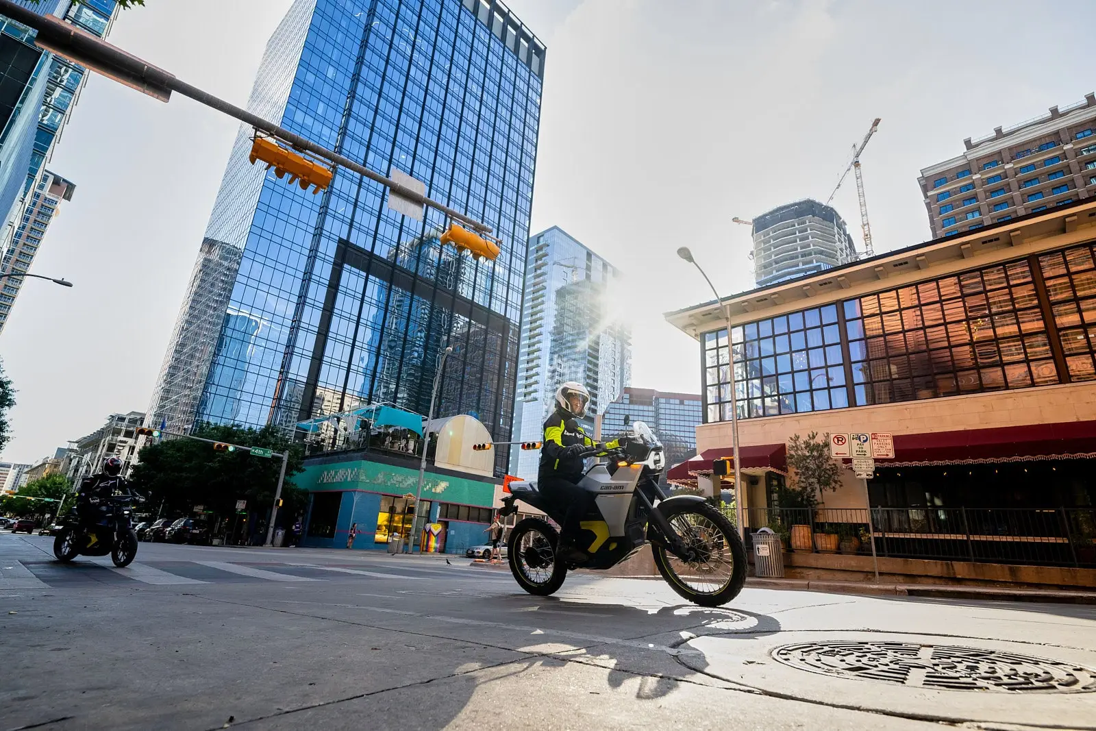 A motorcyclist on a Pulse rides through a street lined with modern glass skyscrapers, bathed in the golden light of late afternoon.