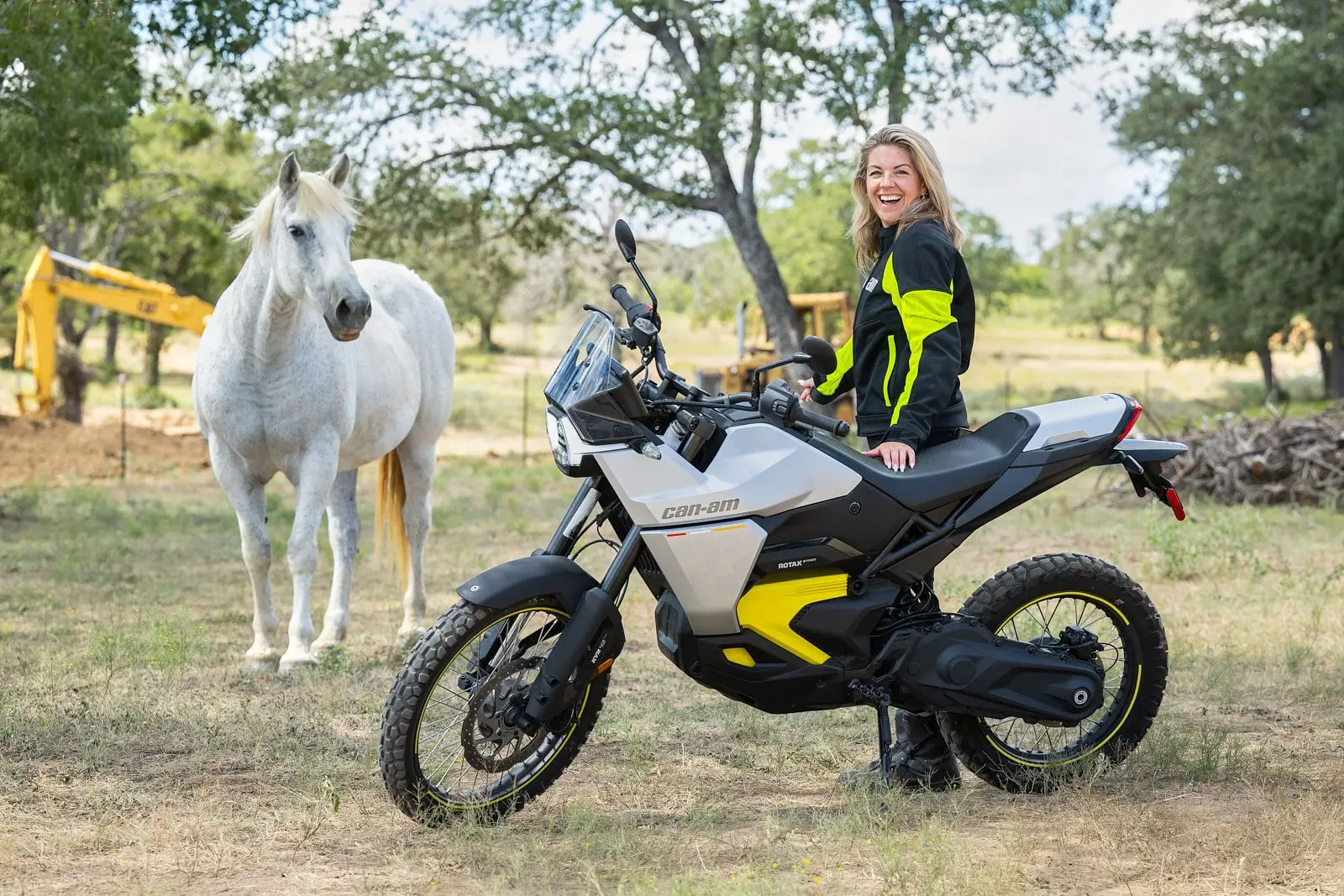 A smiling woman sits on a Can-Am Pulse or Origin electric motorcycle, with a white horse in the background in a natural setting.