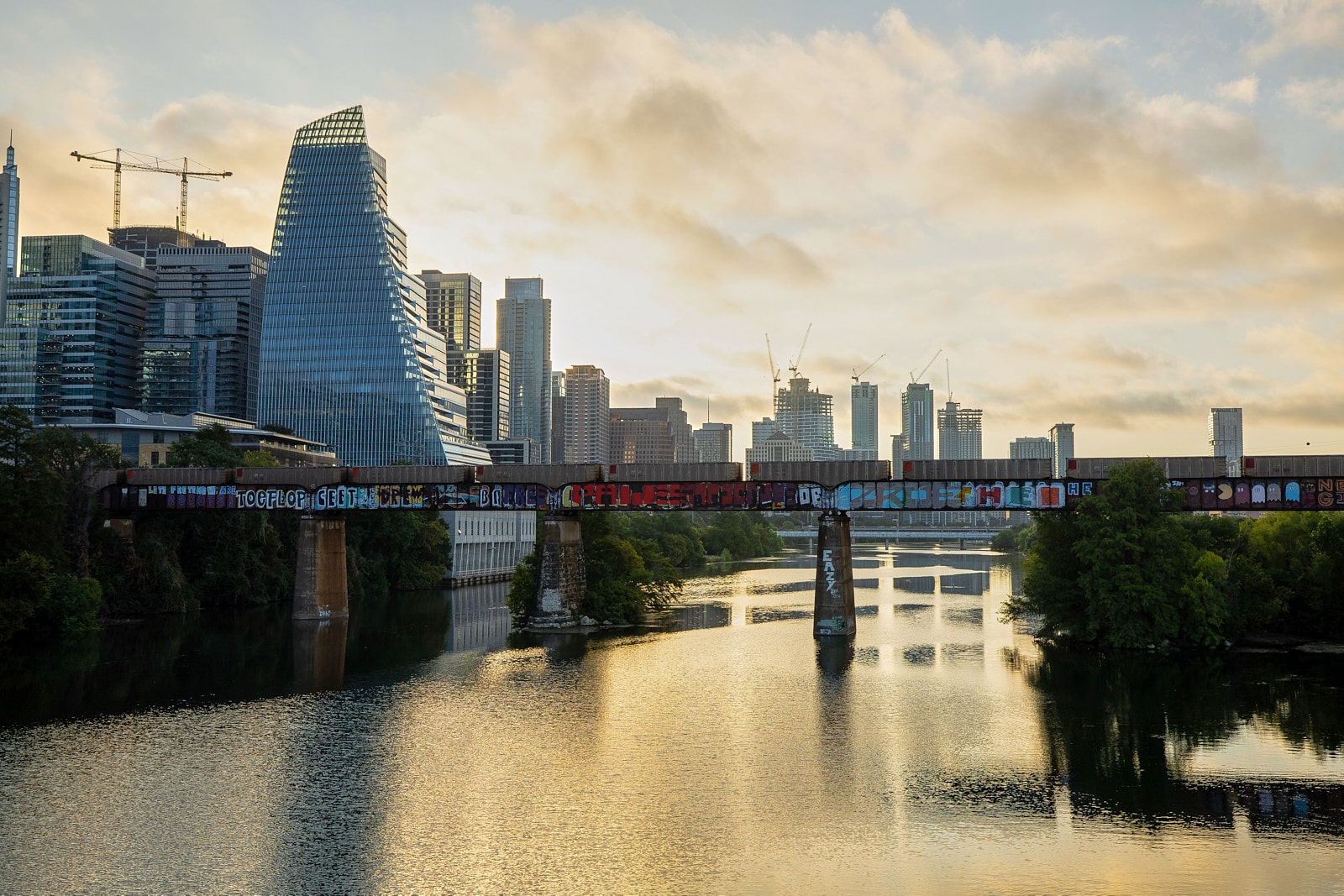 Vue de la ville d’Austin au lever du soleil, avec des gratte-ciel modernes en arrière-plan et un pont ferroviaire couvert de graffitis traversant une rivière calme au premier plan.