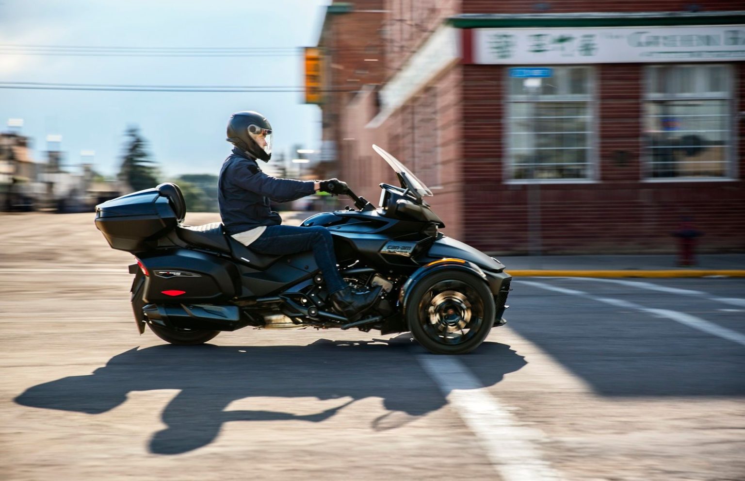 A helmeted biker drives a black trike through a city street, with a brick building in the background.