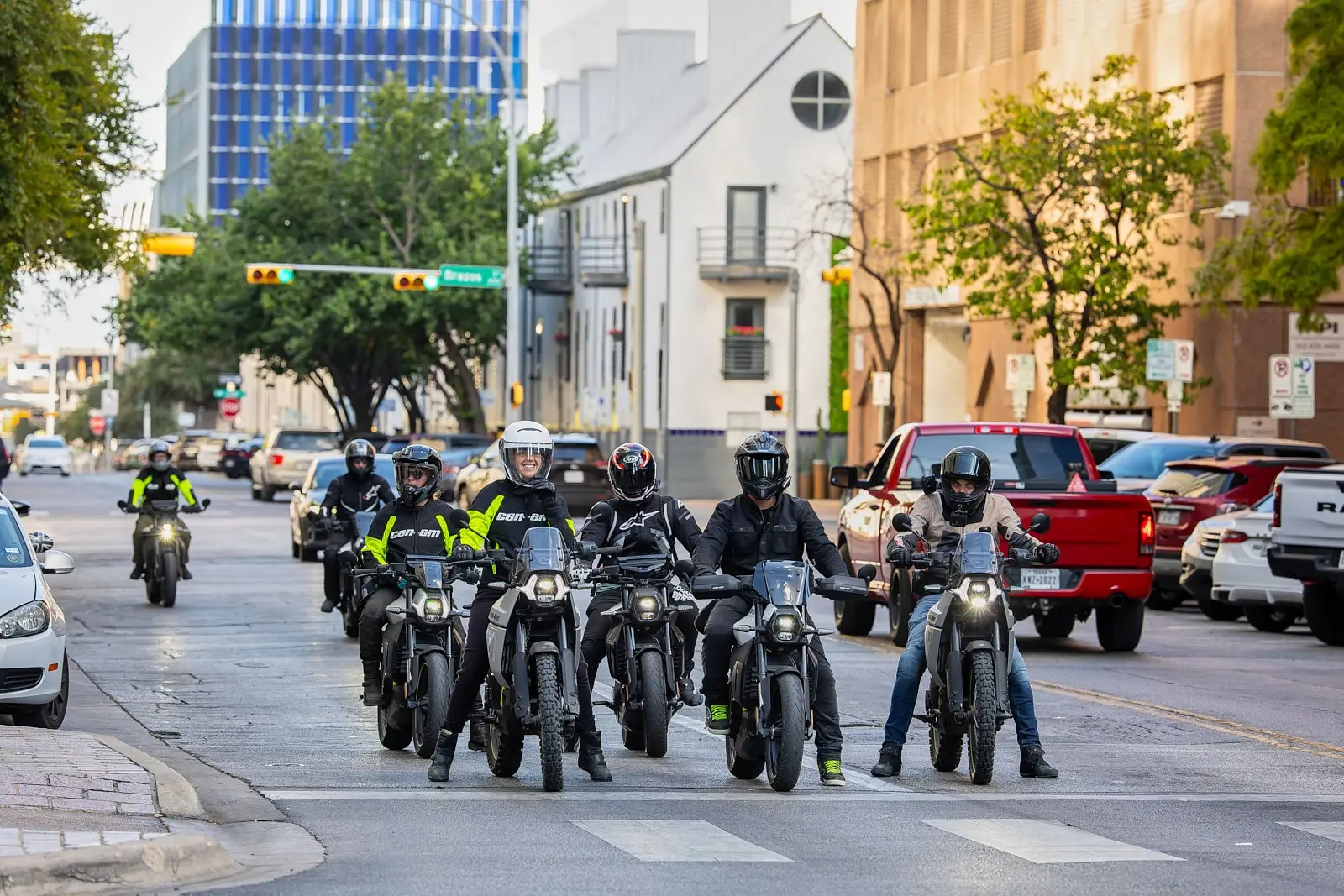 A group of riders on Can-Am electric motorcycles lined up at a traffic light on an urban street.